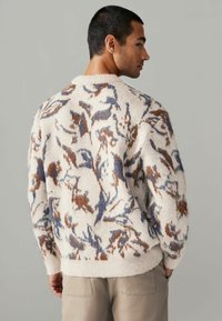 Knit sweater in cream with blue and brown floral patterns, featuring a round neck and ribbed cuffs, visible texture and relaxed fit.