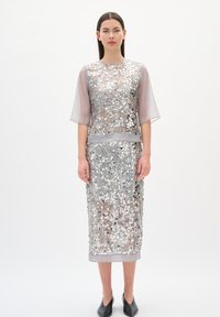 Frau in einem silbernen Paillettenkleid mittlerer Länge mit durchsichtigen ellbogenlangen Ärmeln und schwarzen spitz zulaufenden Schuhen, die vor einem weißen Hintergrund steht.