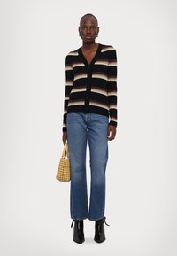 Cardigan negru cu dungi multicolore, closure cu nasturi, asortat cu blugi de culoare albastră deschisă, cizme negre până la gleznă și o geantă țesută galbenă.