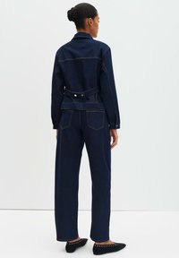 Giacca e pantaloni in denim blu scuro, caratterizzati da una vita aderente con dettaglio fibbia, cuciture visibili e taglio a gamba dritta. Scarpe basse nere.