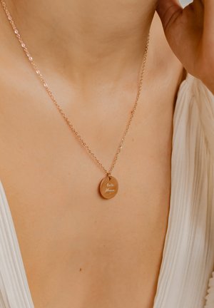 Gold-tone necklace with a round pendant featuring engraved text. The chain is delicate, complementing the smooth surface of the pendant.