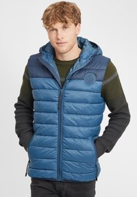 Blå vadderad väst med huva och dragkedja, med en strukturerad övre del och en slät nedre del. Inkluderar sidofickor och en logotyp patch.