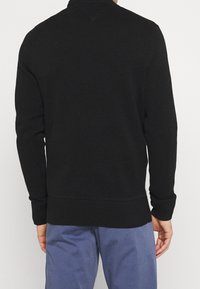 Mann in einem schlichten schwarzen Langarmpullover und blauer Hose, von hinten gesehen, steht vor einem weißen Hintergrund.