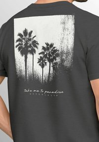 Graues Baumwoll-T-Shirt mit einem weißen Grafikdesign von Palmen und dem Text "Bring mich ins Paradies" darunter, zentriert auf der Rückseite.