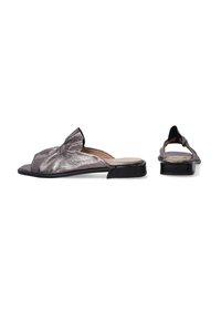 Metallisch silberne Slip-On Sandalen mit einem verdrehten Oberdesign, einer flachen schwarzen Sohle und einer offenen Rückseite für leichtes An- und Ausziehen. Glatte Textur überall.
