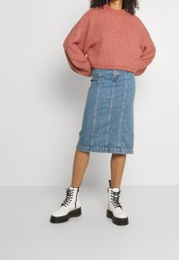 Kvinna i en rosa grovstickad tröja, knälång blå denimkjol och vita snörstövlar med svarta sulor som står mot en enfärgad bakgrund.