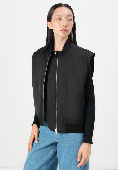 Giacca nera senza maniche con un interno trapuntato, colletto alto e chiusura con zip. Presenta polsini a coste e tasche laterali. Indossata sopra un maglione nero.