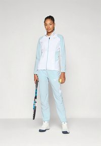 Casaco desportivo azul claro com detalhes brancos e fecho de correr, combinado com calças correspondentes. Modelo segura uma raquete de ténis e uma bola.