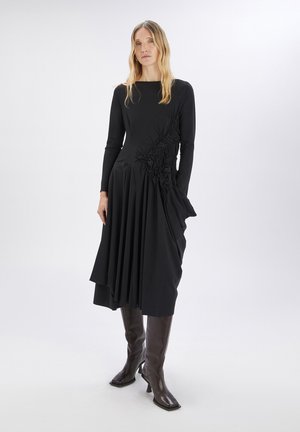 Vestido negro de manga larga con detalles florales texturizados en un lado, falda asimétrica con pliegues y escote redondo. Se lleva con botas hasta la rodilla.
