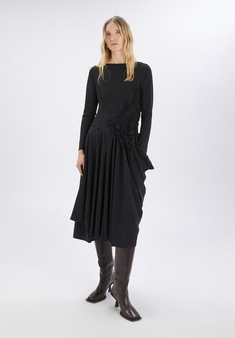 Vestido negro de manga larga con detalles florales texturizados en un lado, falda asimétrica con pliegues y escote redondo. Se lleva con botas hasta la rodilla.