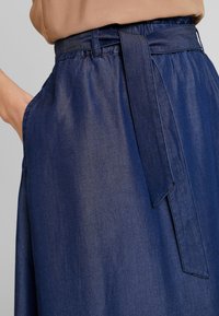 Lähikuvassa henkilö, jolla on tummansininen farkkuhame ja siihen sopiva vyö solmittuna vyötärölle, ja käsi taskussa.