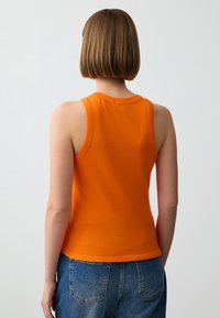 Top arancione senza maniche realizzato in cotone elastico. Presenta un collo rotondo e una forma aderente, abbinato a jeans in denim a vita alta.