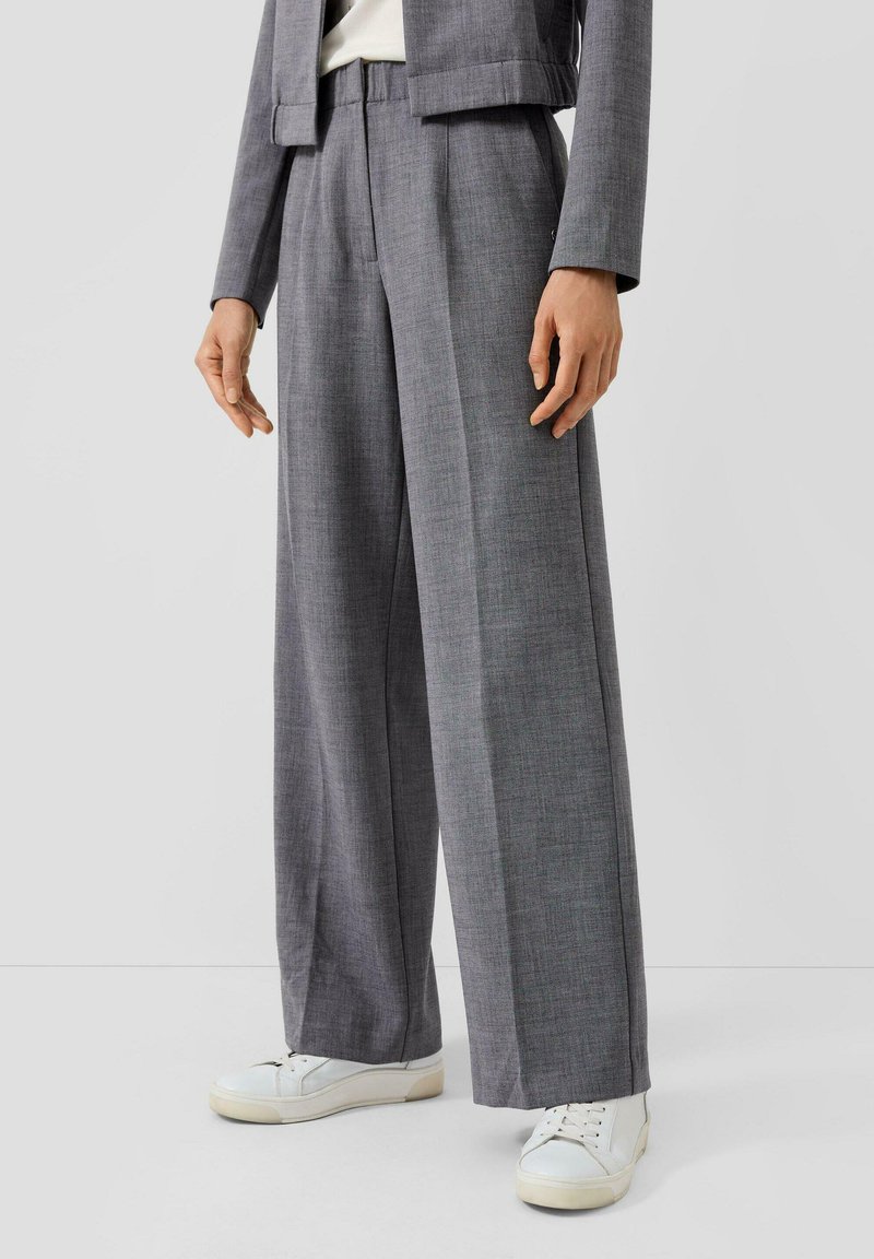 Osoba oblečená v šedých širokých kalhotách a odpovídajícím šedém blejzru, doplněná bílými teniskami, stojící před jednobarevným pozadím.