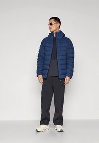 Navy puffer jacket with horizontal stitching, high collar, zip closure, worn over a dark top and paired with dark pants and light sneakers.