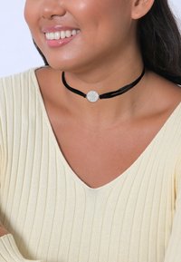 Zwarte fluwelen choker met een zilveren ronde hanger, die een gestructureerd oppervlak benadrukt. Gekleed met een lichtgele ribtop.
