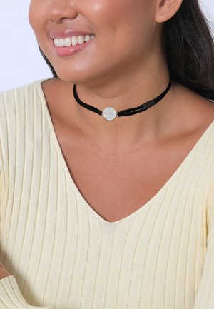 Black velvet choker with a silver circular pendant, highlighting a textured surface. Worn with a light yellow ribbed top.