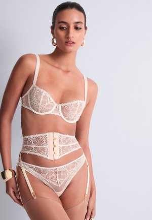 Femme portant un soutien-gorge en dentelle beige, une ceinture porte-jarretelles taille haute, une culotte assortie, des bas transparents, un bracelet en or et des boucles d'oreilles créoles sur un fond uni.