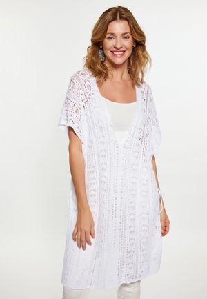 Vrouw met schouderlang lichtbruin haar draagt een witte gehaakte kanten tuniek over een wit topje, glimlachend tegen een effen lichte achtergrond.