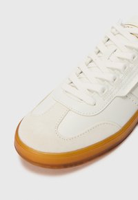 Λευκό αθλητικό παπούτσι με κρεμ λεία σουέτ και λείο δέρμα, στρογγυλή μύτη, λευκές κορδόνες και σόλα από καουτσούκ χρώματος καραμέλας.