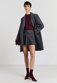 Manteau en laine gris sur un pull côtelé bordeaux, associé à une jupe grise plissée. Les accessoires incluent un sac à main bordeaux assorti et des chaussettes.