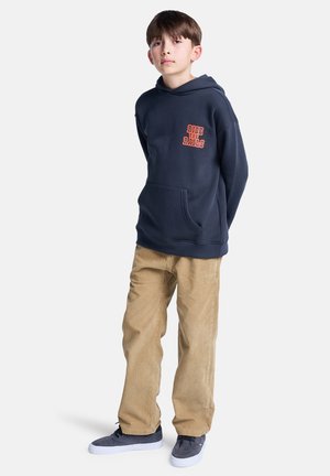 Um jovem rapaz está de pé com confiança, vestindo um hoodie azul-marinho com "RIDE THE RAILS" impresso em laranja, combinado com calças bege e sapatos cinzentos.