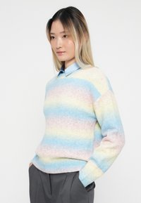 Νεαρός/νεαρή με μακριά ομπρέ μαλλιά, φορώντας ένα παστέλ ριγέ πουλόβερ πάνω από ένα μπλε πουκάμισο και σκούρους γκρίζους παντελόνες, με τα χέρια στις τσέπες, ουδέτερο φόντο.