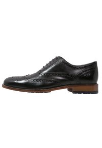 Chaussure brogue pour homme en cuir noir avec design en pointe d'aile, détails perforés, semelle marron et talon bas, présentée en profil latéral.