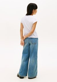 Weißes T-Shirt mit kurzen Ärmeln und gerafften Kanten, kombiniert mit weit geschnittenen blauen Jeans und schwarzen Plateau-Schuhen. Schlichter Hintergrund.