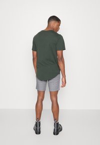 Donkergroene shirt met korte mouwen, grijs geruite shorts, zwarte enkelhoge laarzen met witte stiksels, gezien vanaf de achterkant tegen een effen achtergrond.