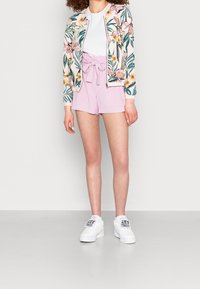 Femme portant des baskets blanches avec des patchs floraux, un short violet clair noué à la taille, un t-shirt blanc et une veste à motif floral.