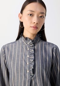 Denimhemd mit vertikalen Streifen, Rüschenausschnitt und Knopfverschluss. Verfügt über hellblaue und dunkelblaue Nuancen mit subtiler Textur.