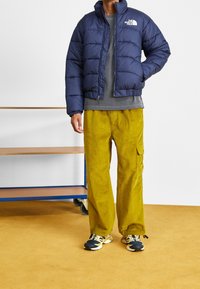 Marinblå puffjacka med hög krage och logotyp, grå skjorta, olivgröna cargobyxor och löparskor med blå och gula accenter.