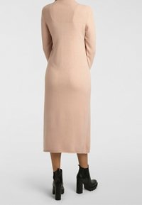 Robe longue moulante en tissu rose doux avec un col côtelé. Présente une texture lisse et s'étend jusqu'au milieu du mollet. Associée à des bottines noires.