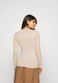 Beige sweater with textured diamond-patterned sleeves and fitted design. Long sleeves and crew neckline enhance the sleek silhouette.