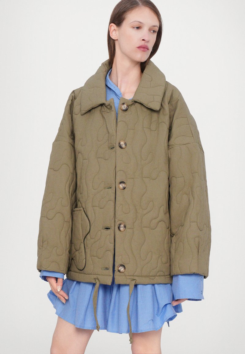 Veste matelassée verte olive avec un grand col, fermeture à bouton, poche avant et ourlet à cordon. En dessous, une chemise à carreaux bleue est visible.