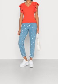 Punainen ribattu V-paitapusero, jossa on röyhelömanchetit, yhdistettynä vaaleansinihin farkkuihin, joissa on valkoisia päivänkakkara-ompeluja ja käännetyt lahkeet, viimeisteltynä valkoisilla lenkkikengillä.