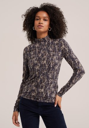 Femme aux cheveux bouclés portant des boucles d’oreilles créoles, un haut à manches longues à col haut et à motifs, et un pantalon foncé sur un fond uni.