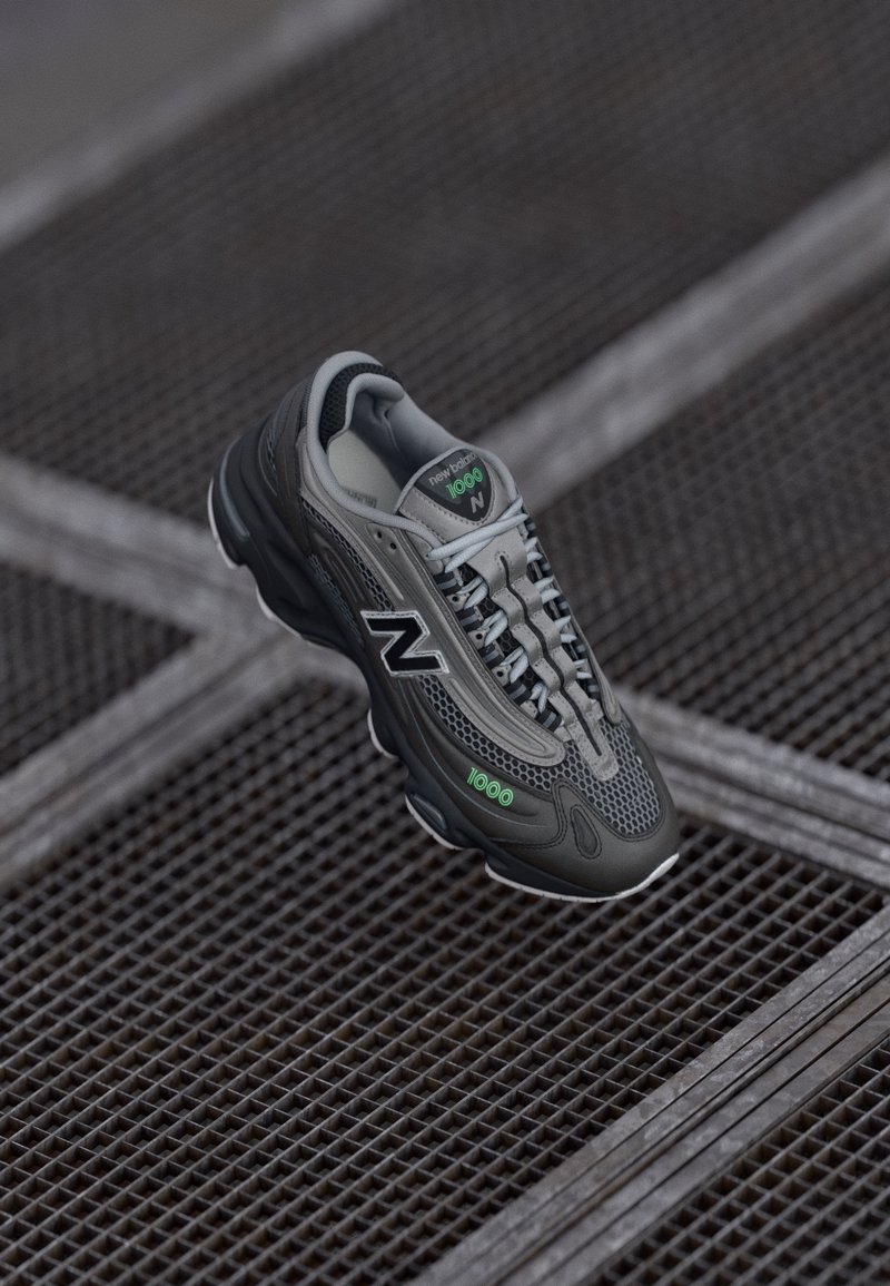 Baskets New Balance 1000 gris foncé et noir avec lacets gris clair, texte vert "1000" et détails en mesh sur une surface en grille métallique.