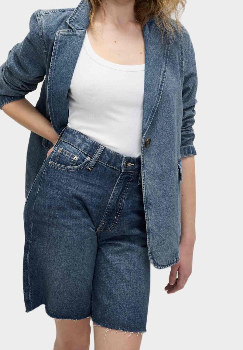 Femme portant un débardeur blanc, un blazer en jean oversize et un short en jean foncé taille haute avec ourlet brut, debout, une main sur la hanche.