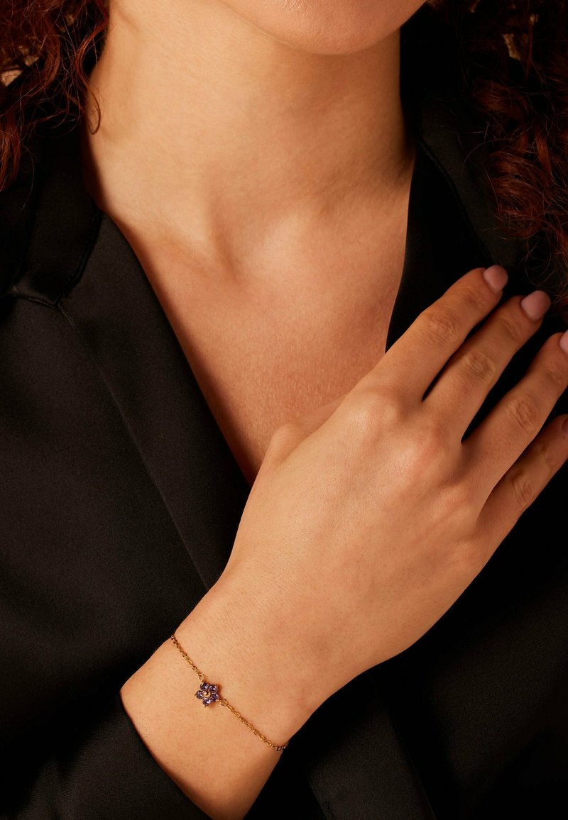 Gouden kettingarmband met een paars bloemenmotief en kleinere kralen. De hand van het model rust op een zwarte blouse.