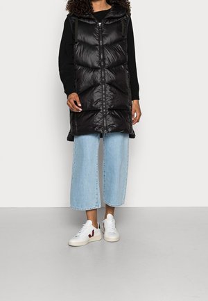 Osoba ubrana w czarną bezrękawnik puchowy na czarną bluzę z długim rękawem, jasnoniebieskie szerokie spodnie typu cropped oraz białe buty sportowe z czerwonym logo.