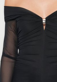 Robe noire à épaules dénudées avec des manches longues transparentes, texture froncée et une accent en métal argenté au niveau du col.