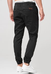 Black jogger pants made of cotton, featuring an elastic waistband, two back pockets, and cuffs at the ankles, paired with white sneakers.