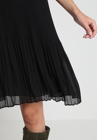 Femme portant une jupe plissée noire et des bottes en daim vert olive, la main détendue le long du corps sur un fond clair uni.