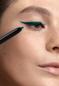 Crayon eye-liner turquoise appliqué sur la paupière supérieure, mettant en valeur un design ailé net. La peau est claire avec une texture naturelle et de légères taches de rousseur.