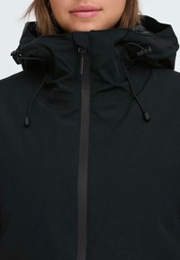 Chaqueta negra con un cuello alto, cordones ajustables y un cierre frontal. Textura de tela suave, diseño minimalista sin patrones visibles.