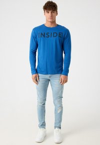 Camiseta de manga larga azul con la palabra "INSIDE" impresa en negro en el pecho, combinada con jeans claros desgastados y zapatillas blancas.