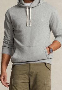 Felpa grigia con cappuccio, nastri e tasca frontale. Presenta un piccolo logo sul petto. Indossata con pantaloni cargo verdi e un orologio.