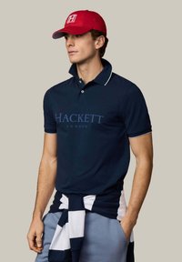 Mladý muž, ktorý nosí námornícky modré polokošeľu značky „Hackett London“, červenú čiapku s logom „H“, pruhovaný sveter zviazaný okolo pása a svetlomodré nohavice.