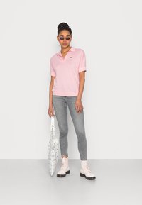 Różowa koszulka polo z krótkim rękawem, szare skinny jeansy i biała torba. Na nogach jasnoróżowe buty wojskowe i okulary przeciwsłoneczne. Zielony detal z logo.